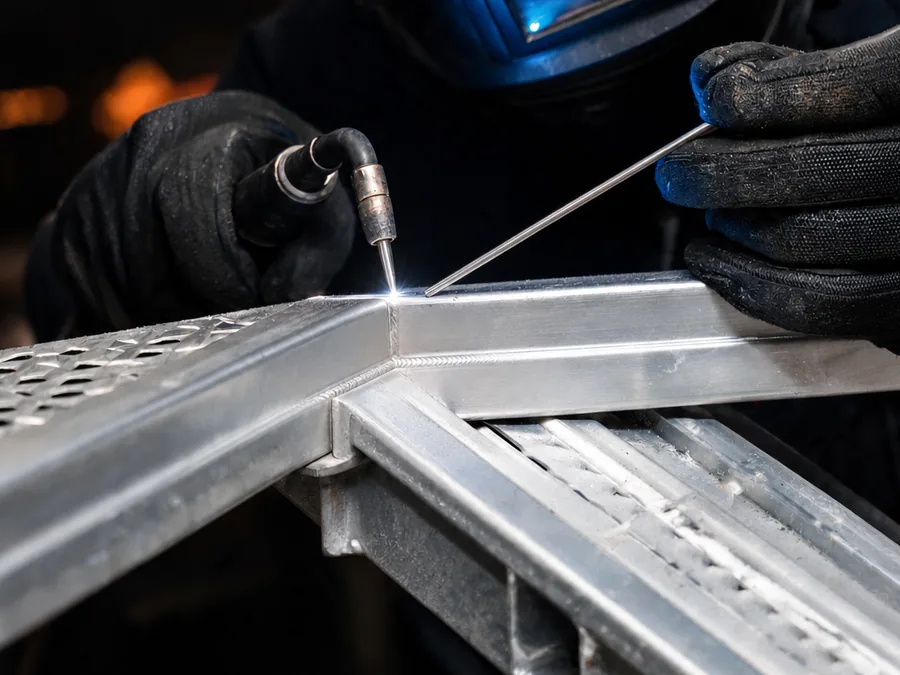 Close-up of a TIG weld repair on a cracked aluminum trailer ramp frame, with tungsten torch and filler rod