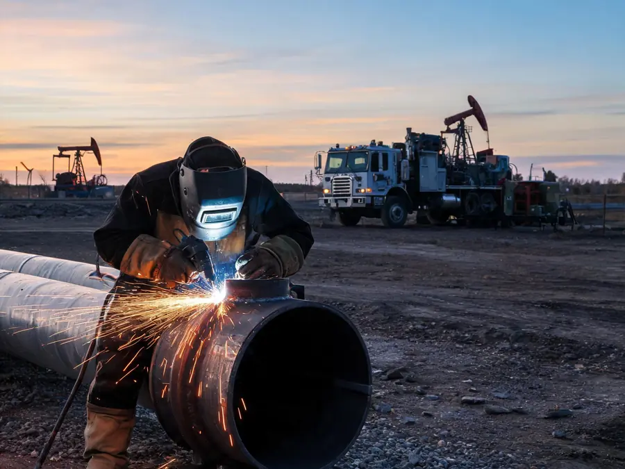Rig welder welding a steel pipe spool at an oilfield site at dusk with a welding truck behind