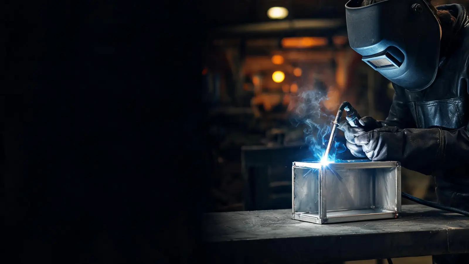 Welder TIG welding an aluminum box frame on a steel table in a dark Calgary fabrication shop, blue arc glow on the aluminum
