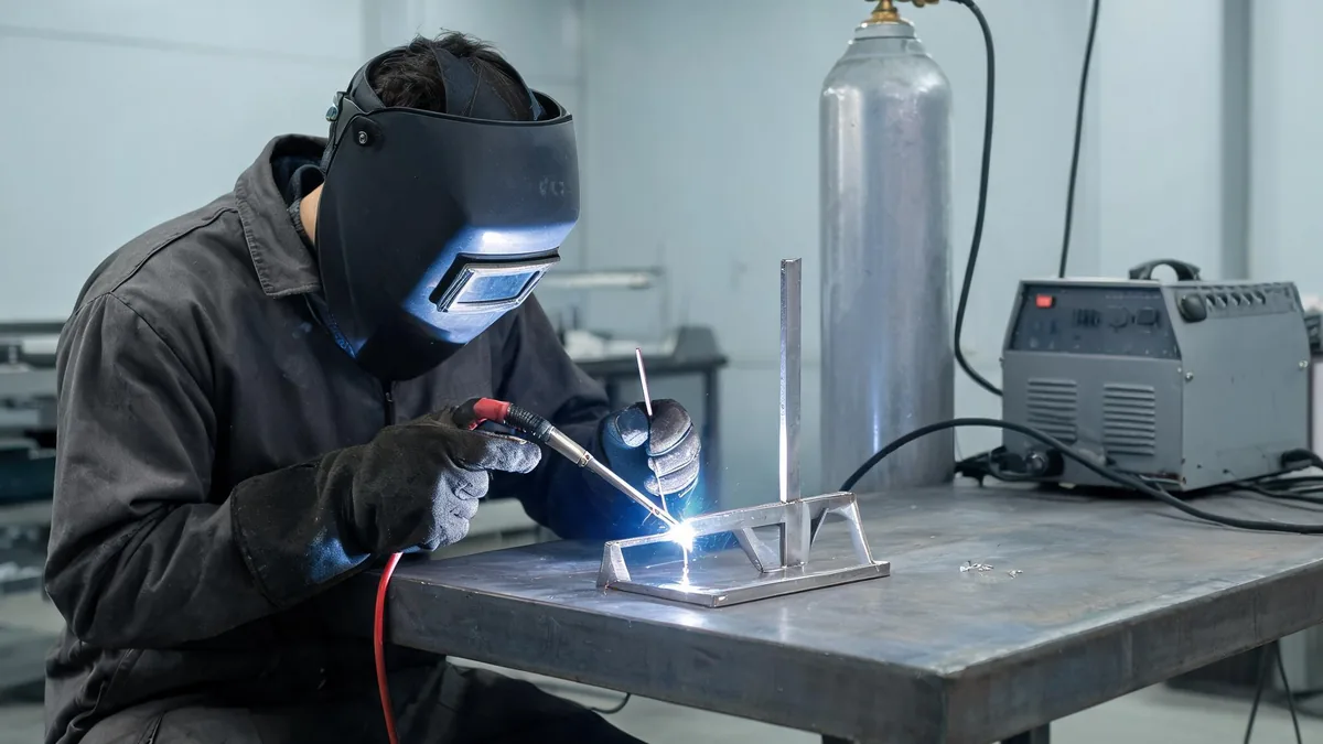 Welder at a bench TIG welding a thin stainless steel bracket while feeding filler rod
