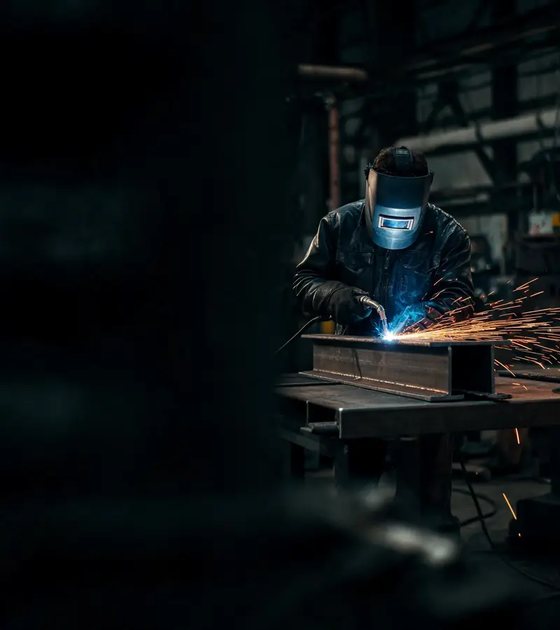 Welder in a helmet MIG welding a steel beam with sparks flying in a dark fabrication shop