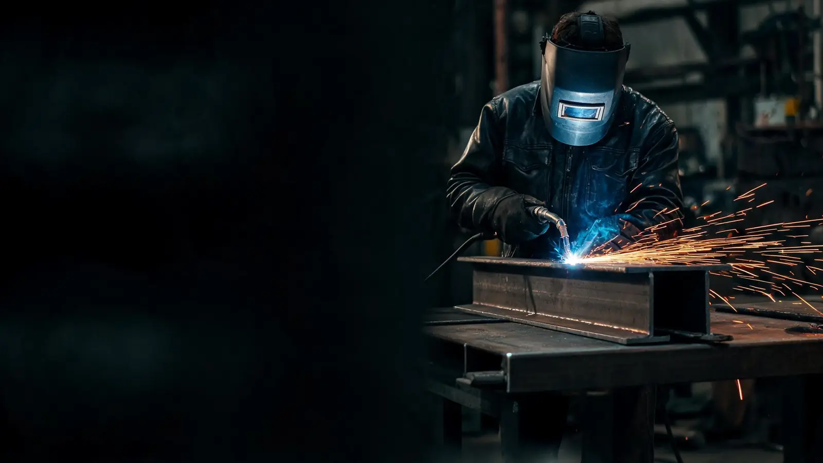 Welder in a helmet MIG welding a steel beam with sparks flying in a dark fabrication shop