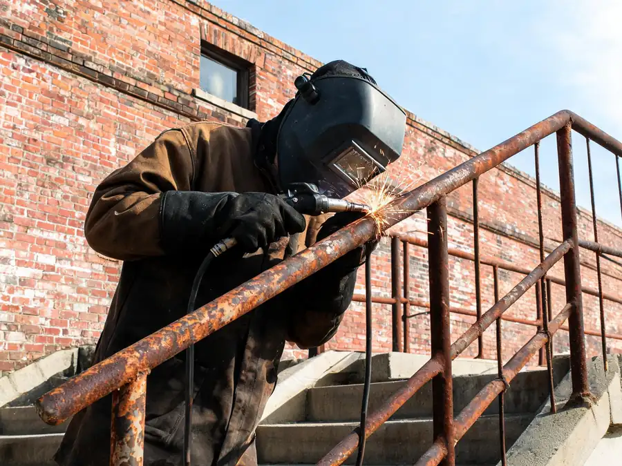 Welder repairing a rusty steel stair railing outside an older brick building